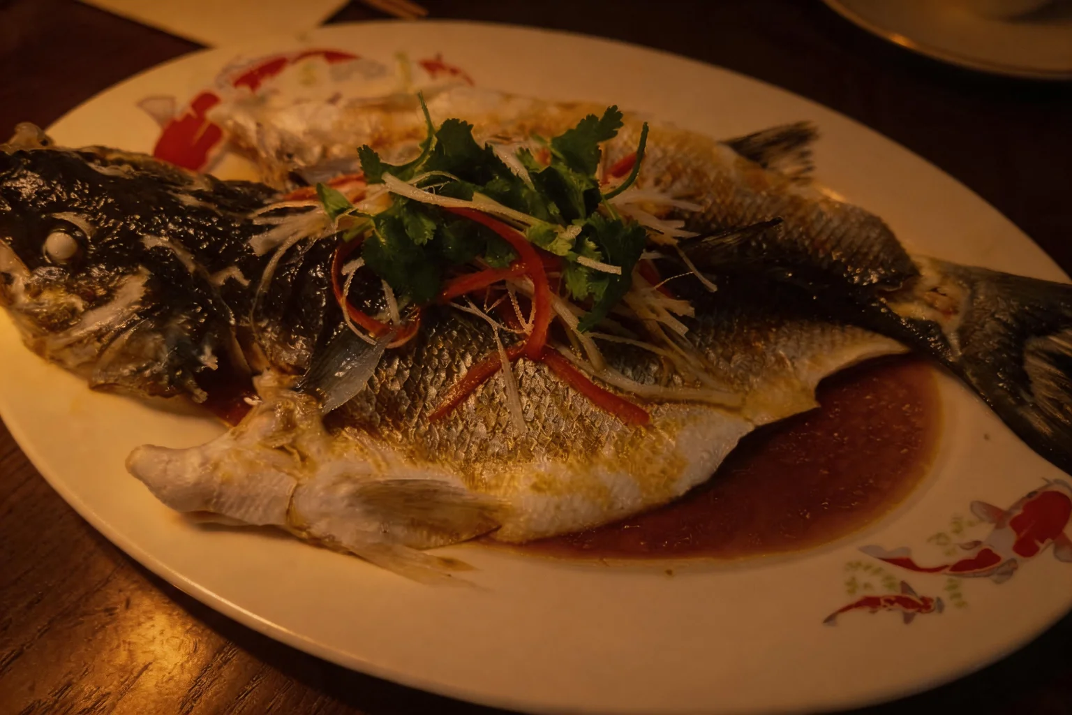 Bar entier cuisiné à la vapeur sur un plat blanc à motifs de carpes, lumière chaude du soir